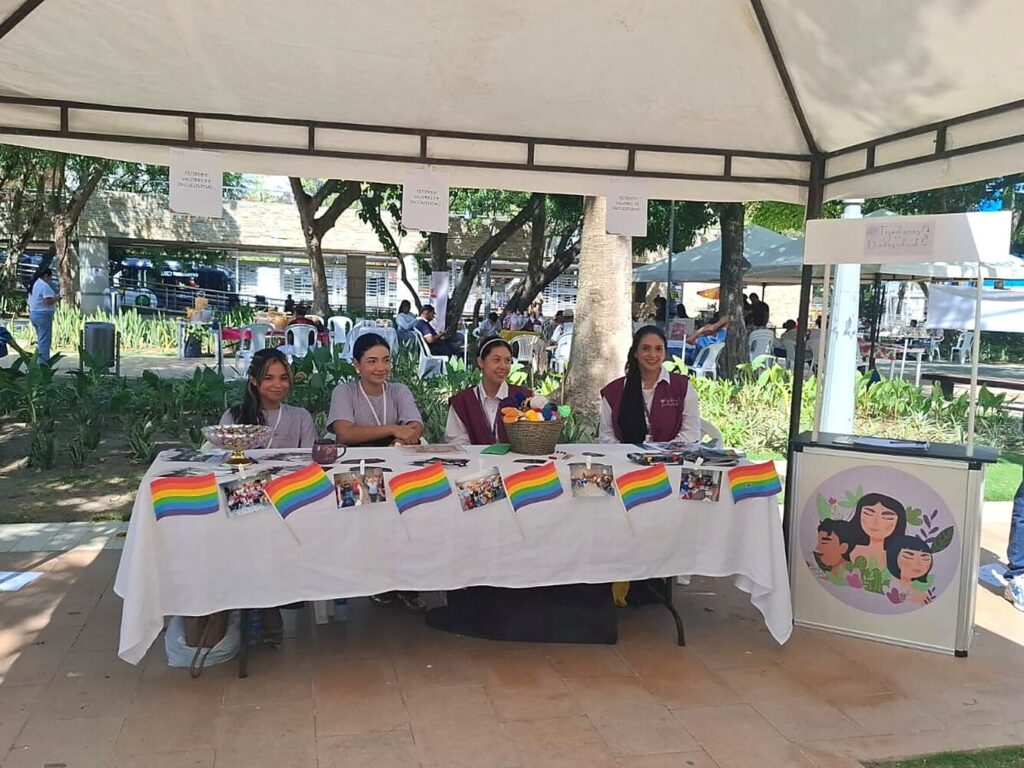 Integrantes de la Fundación atienden un puesto informativo al aire libre, con material de la organización y banderas de la diversidad sobre la mesa.